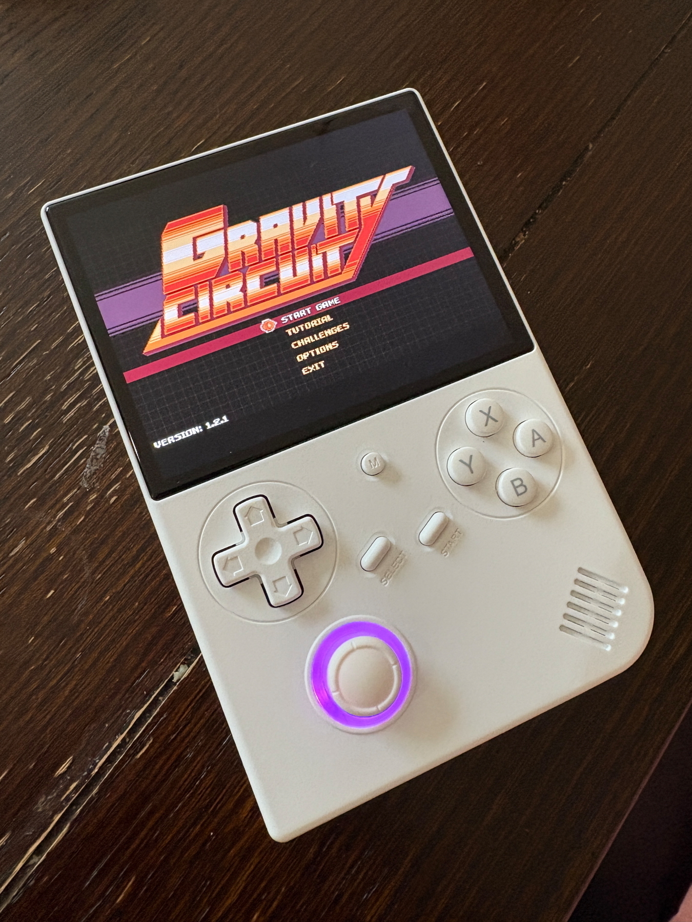 A handheld gaming device with an active screen displaying the “Gravity Circuit” game menu. The device has a D-pad, four buttons labeled X, Y, A, and B, and two smaller buttons labeled “Select” and “Start.