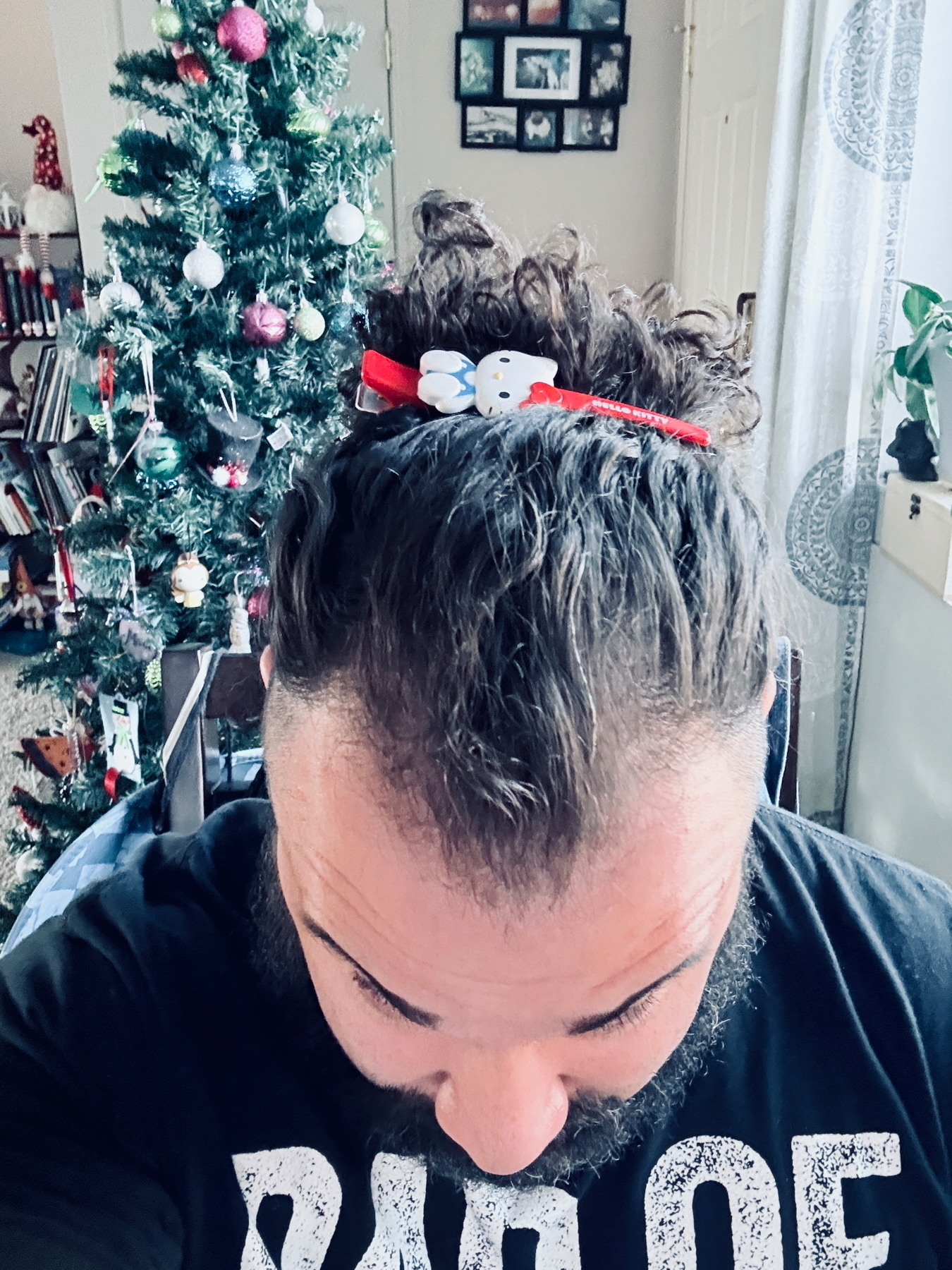 A person with a beard is looking down, wearing a red Hello Kitty headband. In the background, there is a decorated Christmas tree with colorful ornaments and a wall with framed photos.