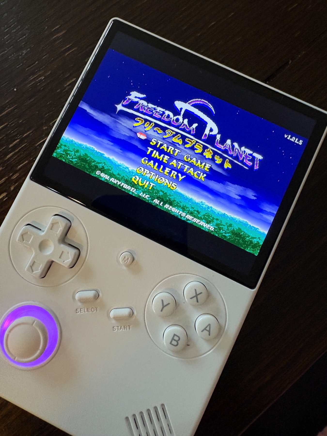 A handheld gaming console displaying the title screen of the game “Freedom Planet.” The console has a white design with a D-pad, buttons labeled A, B, X, Y, a glowing analog stick, and a screen showing menu options like Start Game