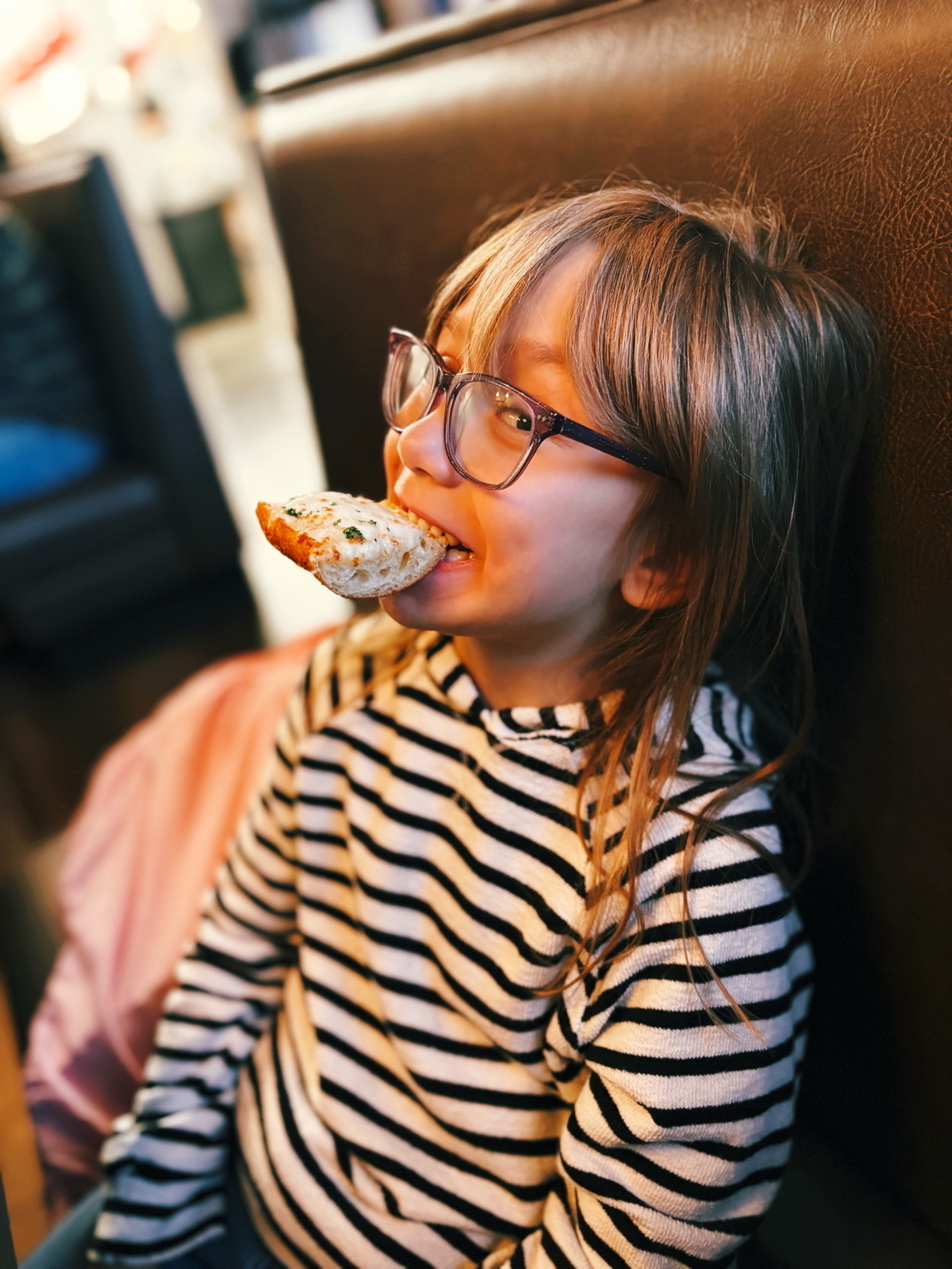 A child with glasses is joyfully biting into a piece of garlic bread. They are wearing a black and white striped shirt and are seated in a cozy setting.