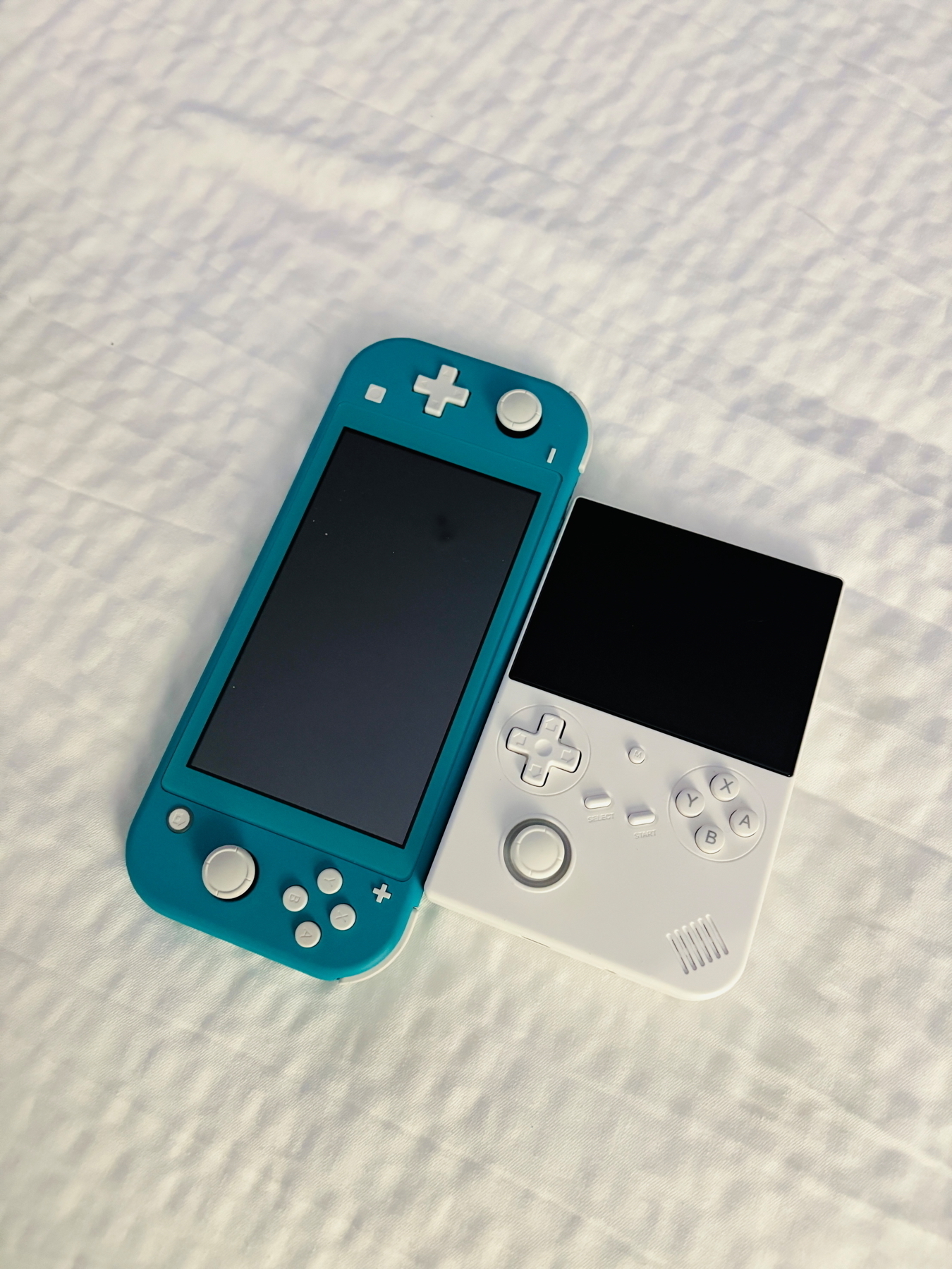 A teal handheld gaming console and a white retro-style handheld console are placed side by side on a white textured surface.