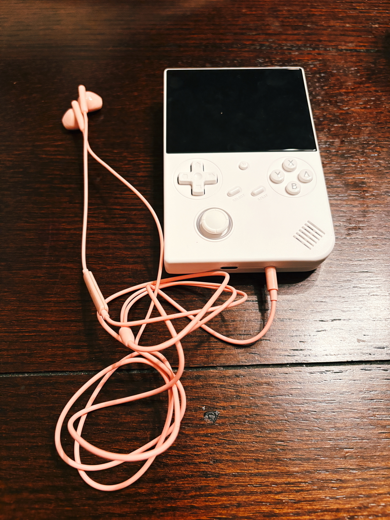 A handheld gaming console with a blank screen and a set of pink earphones resting on a wooden surface.