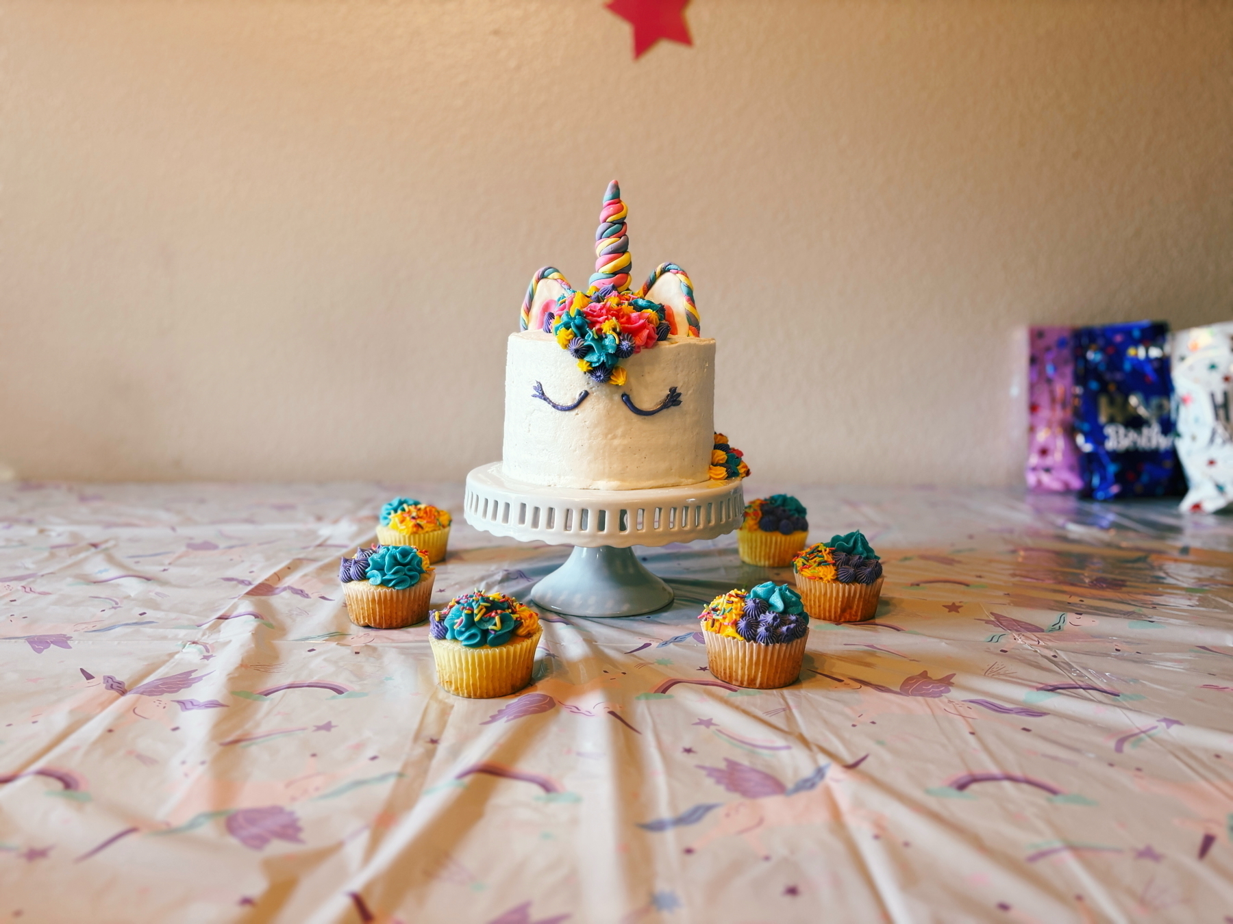 A colorful unicorn-themed cake with a horn and floral decorations on a cake stand, surrounded by six cupcakes with rainbow frosting on a pastel tablecloth. Birthday gift bags are in the background.