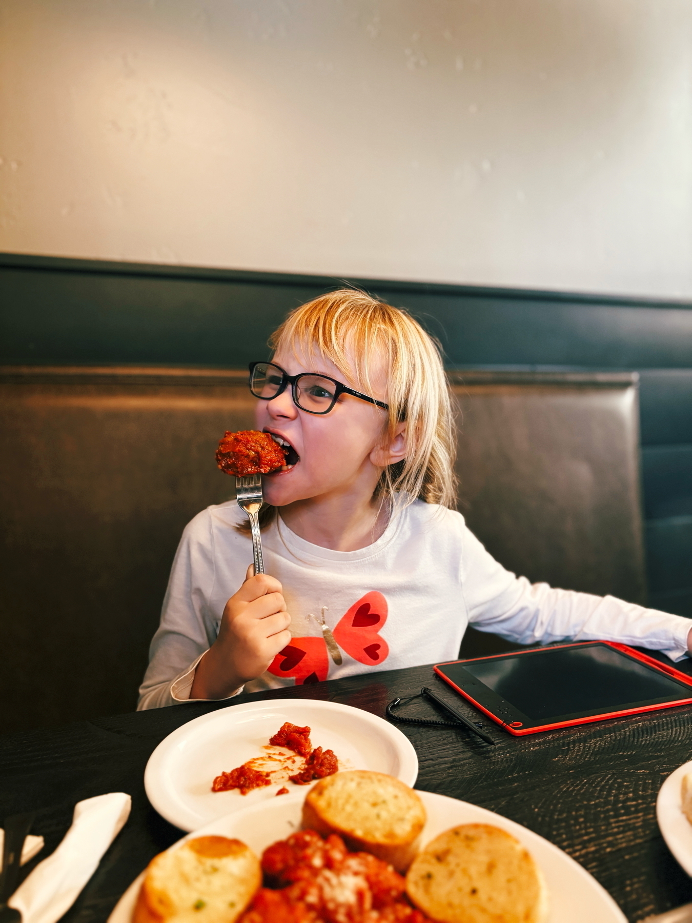 A child with glasses is eating a large meatball on a fork. The child is wearing a white shirt with a red butterfly design. In the foreground, there is a plate with pasta and garlic bread. An electronic tablet is on the table next to