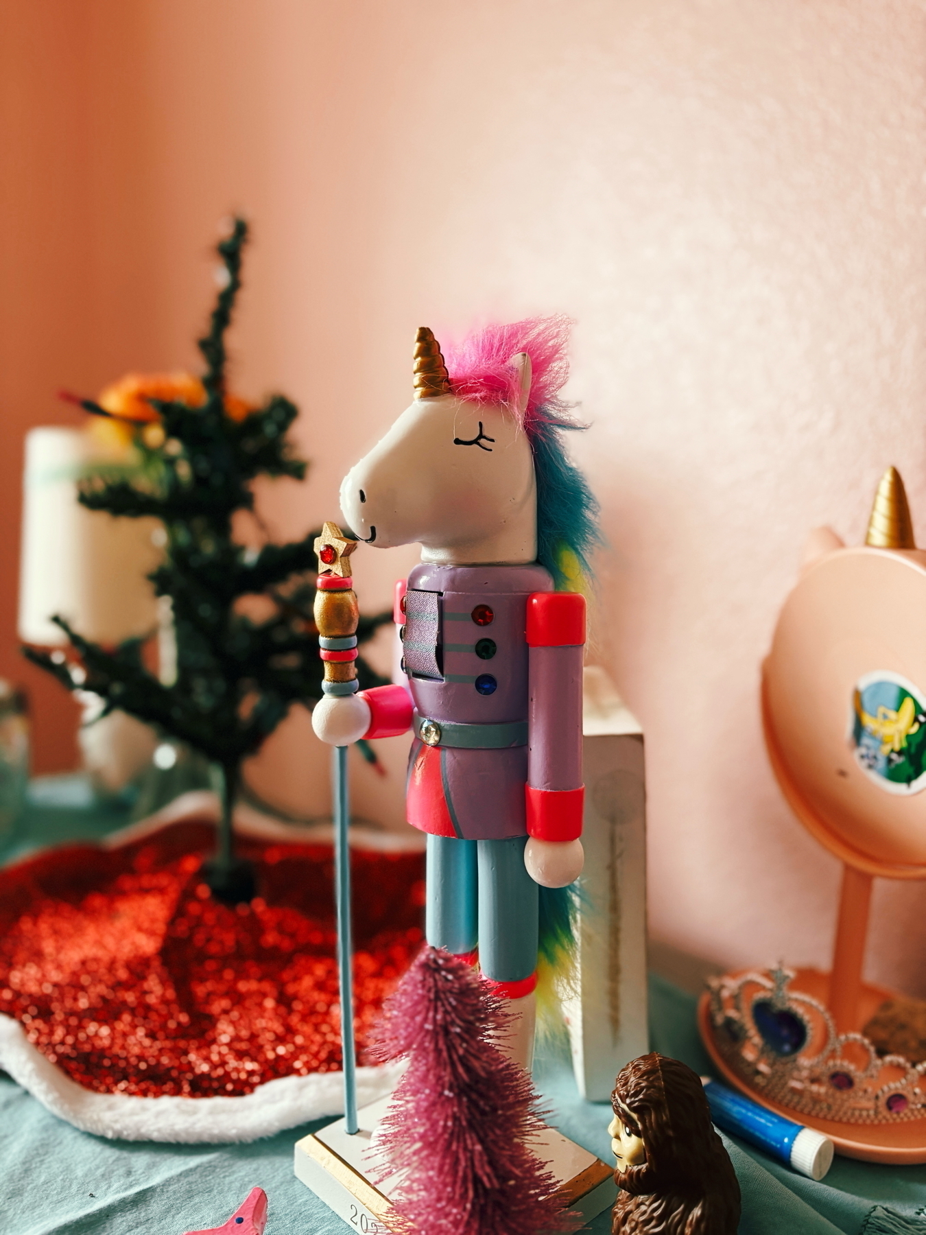 A colorful unicorn nutcracker with a rainbow mane and horn stands on a table. Nearby are small decorative items, including a mini Christmas tree, a toy animal, a decorative crown, and a pink backdrop.