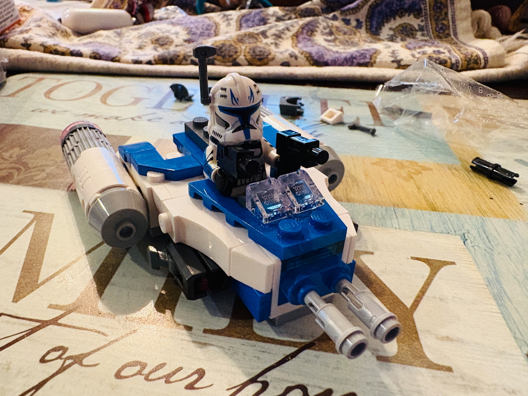 LEGO Star Wars clone trooper minifigure sitting in a small blue and white spaceship with dual cannons, surrounded by additional LEGO pieces on a table with a patterned cloth in the background.