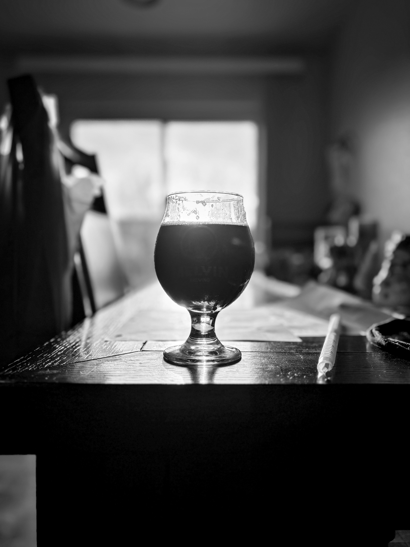 A black and white photo of a rounded glass containing a dark beverage placed on a wooden table. The background is blurred, with light coming through a window, creating a dramatic contrast. A straw lies beside the glass.