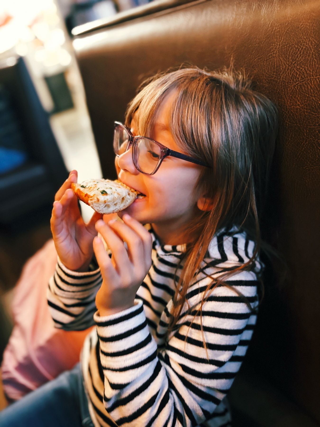 A young girl wearing glasses and a striped sweater is sitting on a bench, enjoying a slice of bread.