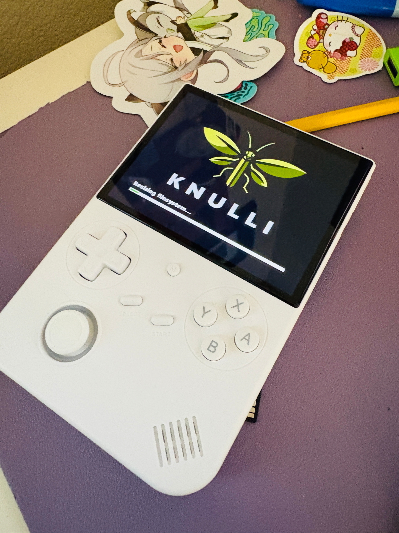 A white handheld gaming device with an off-center screen displaying a logo and text “KNULLI” with a green insect design. The device has Y, X, A, B buttons, a directional pad, and a start/select button. Surrounding