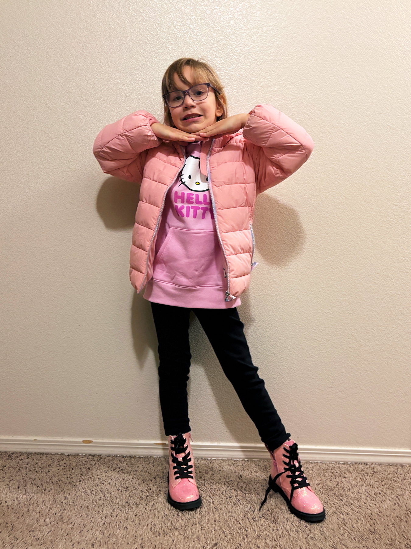 A child stands against a wall, wearing glasses, a pink puffer jacket, a Hello Kitty hoodie, black leggings, and pink shiny boots. The child has their hands under their chin, striking a playful pose.