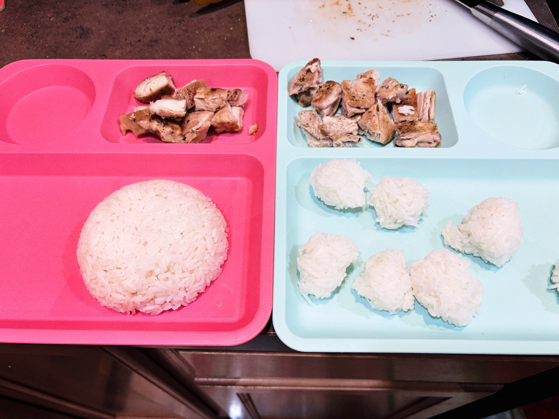 Two colorful plates with sections. The pink plate contains a mound of rice and pieces of cooked chicken. The blue plate has several small rice balls and more pieces of cooked chicken.